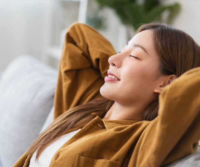 A woman with long hair relaxes on a couch, leaning back with her hands behind her head and eyes closed, smiling peacefully—perhaps unwinding after overseeing a roofing process. She is wearing a brown shirt. The background is softly blurred.