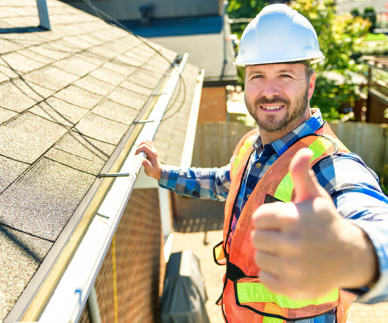 A construction worker wearing a hard hat and safety vest stands on a ladder next to a roof gutter, smiling and giving a thumbs-up to the camera, showcasing professional roofing Ottawa services on a sunny day.