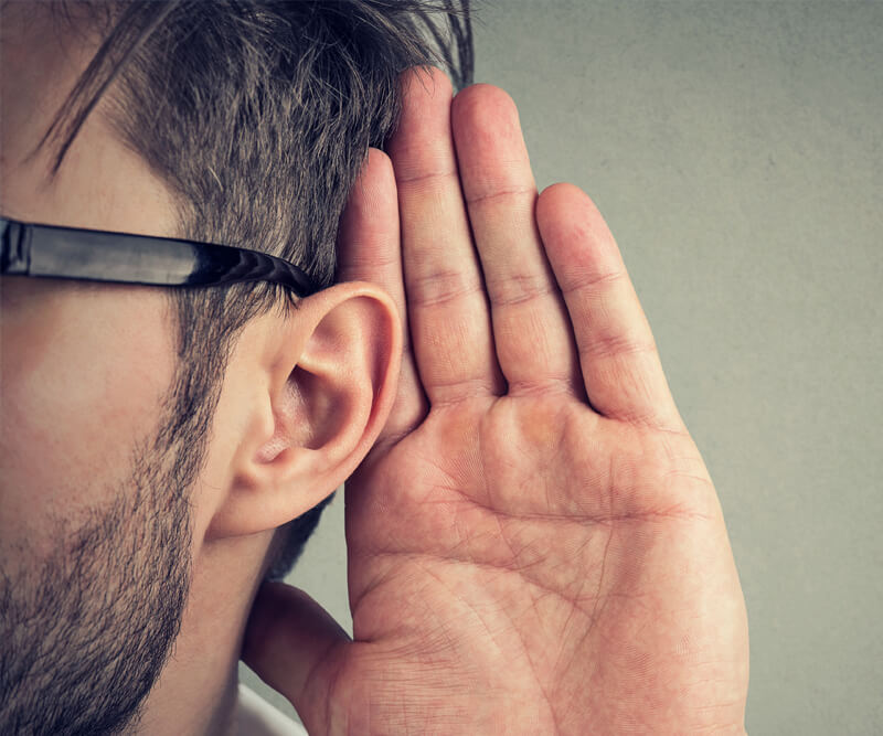 A person wearing glasses holds their hand up to their ear, appearing to listen closely. Only the side of the persons face and the hand are visible, with a neutral background.