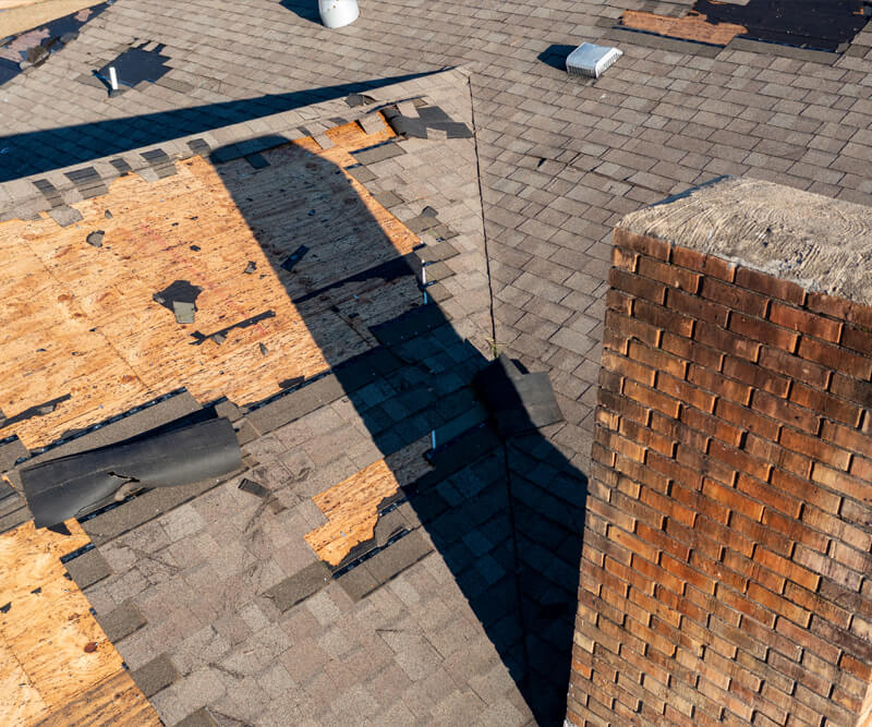 A roof with missing and damaged shingles exposes the wooden decking beneath. A brick chimney stands on the right, and scattered roofing materials suggest recent storm damage or repair work—an ideal situation to review your roofing insurance coverage.