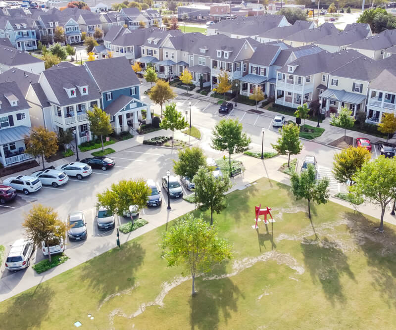 Aerial view of a suburban neighborhood with rows of houses featuring Stittsville roofing, parked cars along the street, and a small park area with green grass, trees, and a red playground structure.