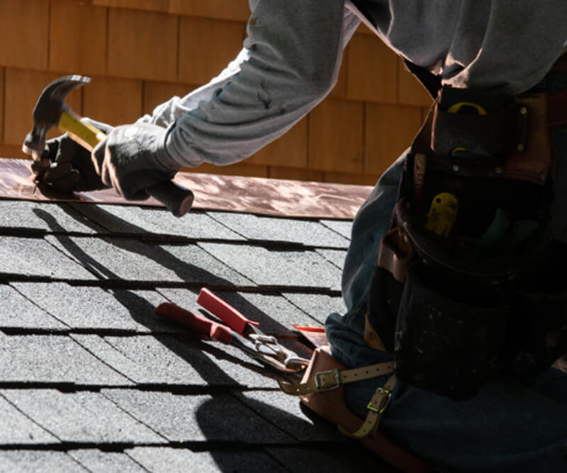 A worker wearing a tool belt uses a hammer to install or repair asphalt shingles on a roof beneath sunlight. The persons face is not visible, capturing the dedication seen in Riverside South roofing projects.