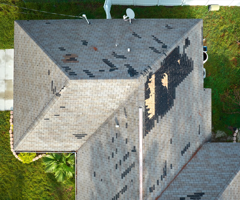 Aerial view of a house roof with missing and damaged shingles, revealing exposed areas in need of roof repairs Riverside South. The surrounding yard is green with grass and a few plants visible.