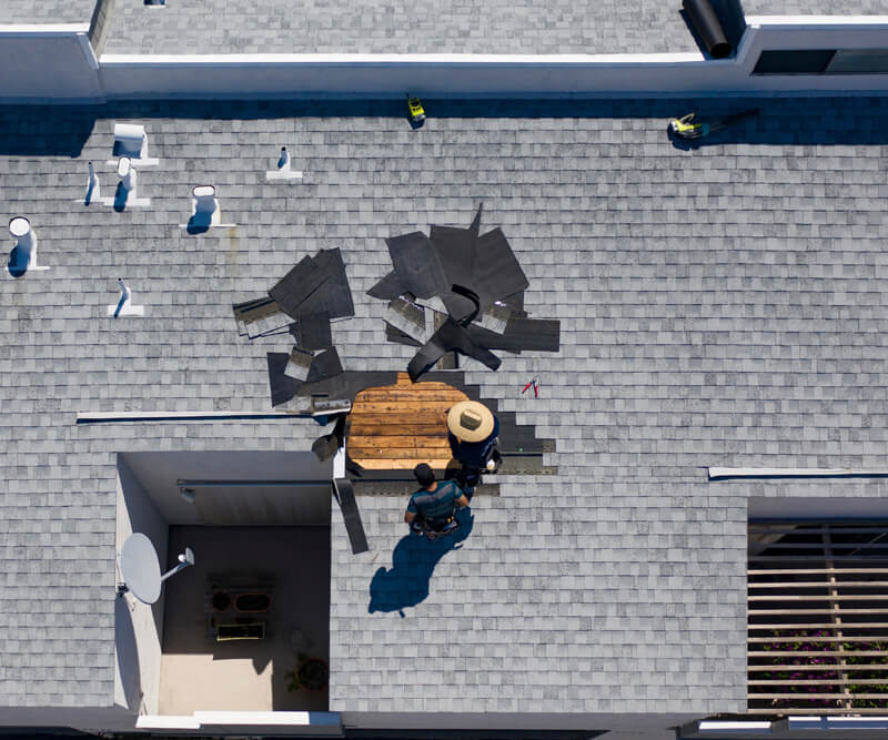 A person in a hat works on roof damage repairs, surrounded by scattered roofing materials and tools. The image, taken from above, shows a section of exposed wood where shingles have been removed.