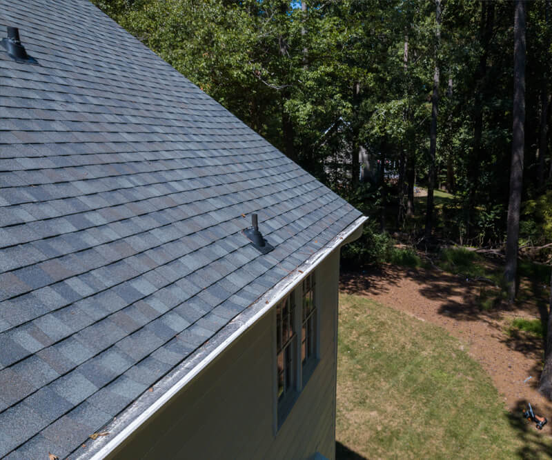 A roof features gray asphalt shingles and two vent pipes, viewed from above, with a grassy yard and trees in the background on a sunny day in an Orleans neighbourhood.