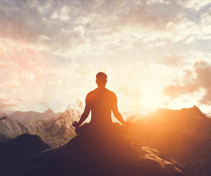 A person sits cross-legged on a mountaintop, meditating at sunrise or sunset, embracing peace of mind as the mountains and a colorful sky create a serene backdrop.