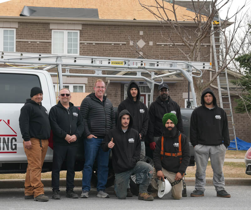 The Firon Roofing team in Ottawa, eight men in work attire, pose in front of a van with a ladder on top. Some stand while others kneel outdoors near houses, dressed in hoodies and jackets.