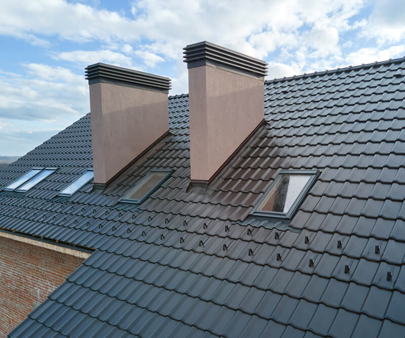 A modern sloped roof with dark gray tiles, two large rectangular chimneys, and several skylight windows—ideal for those considering skylight replacement in Ottawa—stands out under a partly cloudy sky.