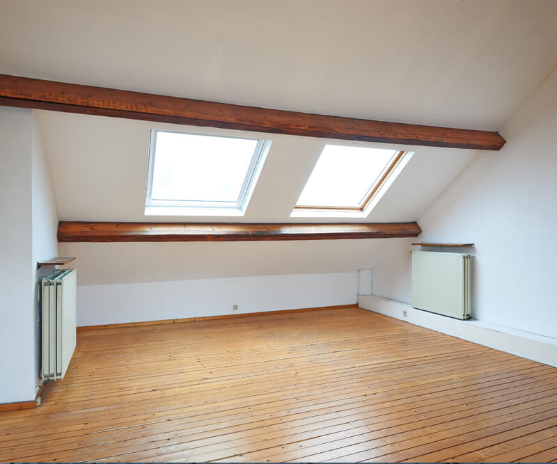 A bright attic room with wooden floors, white walls, exposed wooden beams, and two large skylights letting in natural light—ideal for a future skylight replacement. Two radiators sit on opposite walls beneath the sloped ceiling.