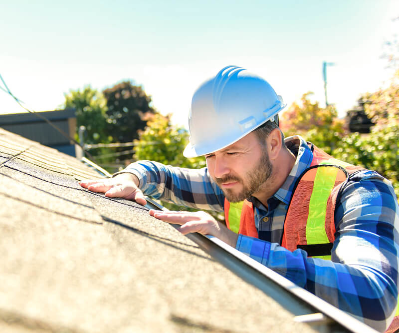 A construction worker wearing a white hard hat, safety vest, and plaid shirt inspects a residential roof under bright sunlight, checking the shingles for quality to ensure the use of the best roofing products. Trees and sky are visible in the background.