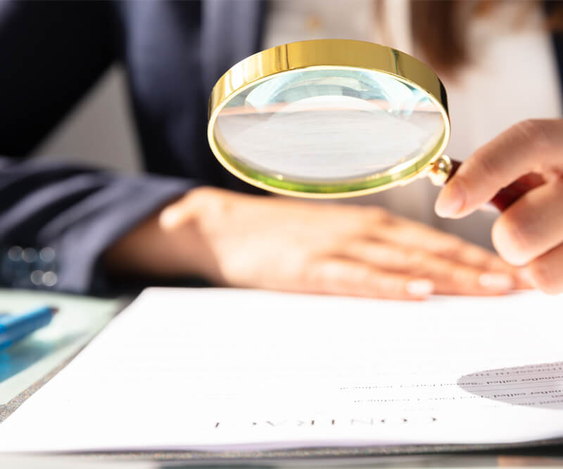 A person holding a magnifying glass over a document, closely examining the fine-print warranty. The individual is wearing a dark suit, and only their hands and partial torso are visible.