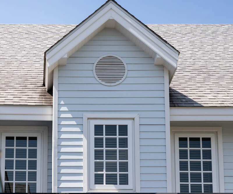 Close-up of a light blue house exterior with white trim, featuring a gable roof, a round attic vent, and two windows under a clear blue sky—ideal for showcasing roof inspection Ottawa services.