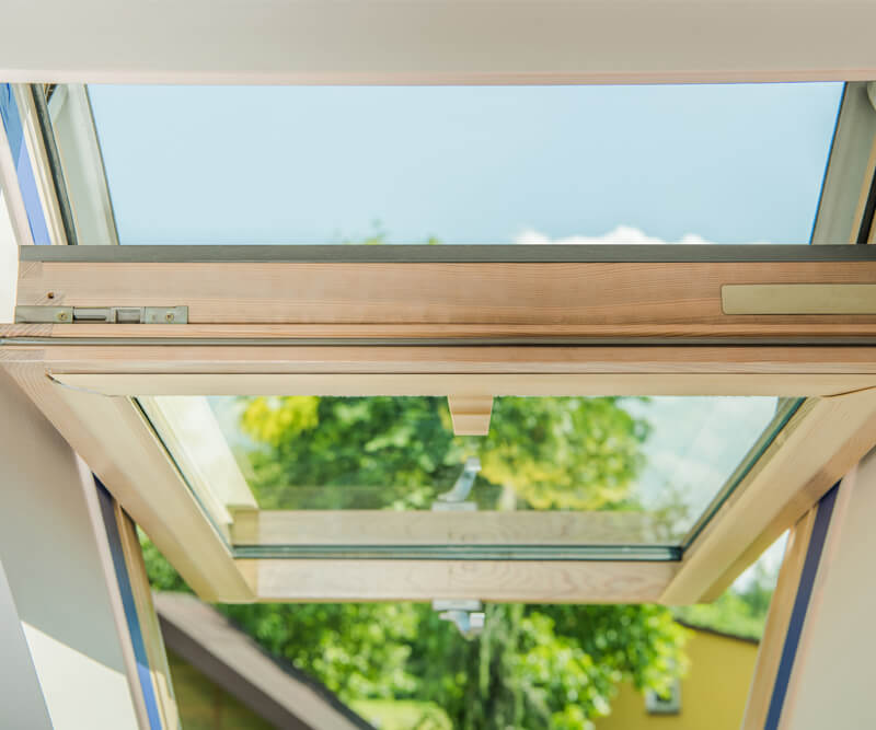 Open skylight window in a slanted roof, showing a clear blue sky and green trees outside. Sunlight streams through the glass, illuminating the wooden frame—a perfect example of quality skylights Nepean residents enjoy.