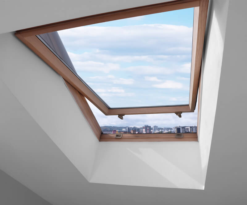 A skylight Nepean window set in a sloped white ceiling is open, revealing a view of a blue sky with clouds and a city skyline in the distance. The window has a wooden frame.