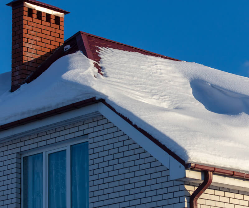 A house with white brick walls and a red roof covered in thick snow, with a red brick chimney and blue sky in the background—perfectly capturing a scene before roof snow removal Ottawa services are needed. Snow piles up on the sloped roof above a window.