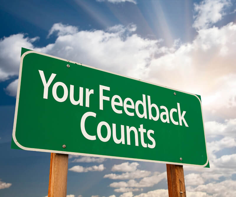 A green sign with white text that reads “Your Feedback Counts” stands outdoors against a partly cloudy sky with sun rays shining through, reminding customers how Firon Roofing reviews can make a difference.