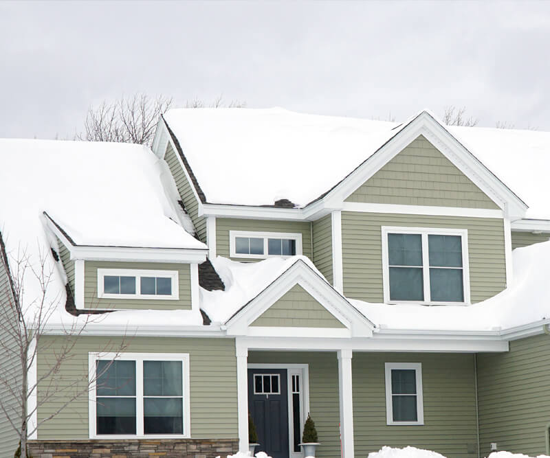 A green two-story house with white trim sits under a cloudy winter sky, its roof and yard blanketed in snow—an ideal candidate for professional snow removal Manotick services.