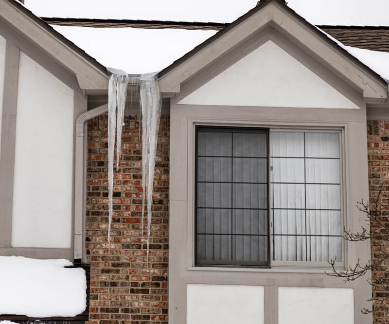 Two large icicles hang from the gutter of a brick house with snow on the roof and ground—an all-too-common sight before ice dam removal in Manotick. A window with closed blinds is visible beneath the thick icicles.