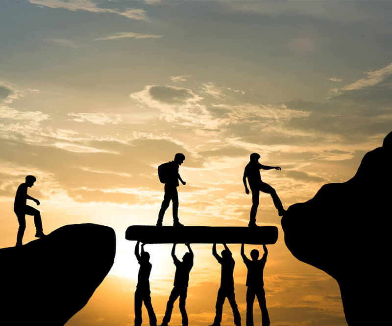 Silhouettes of people at sunset, some holding up a beam to form a bridge while others walk across or prepare to step onto it, symbolizing teamwork and cooperation.