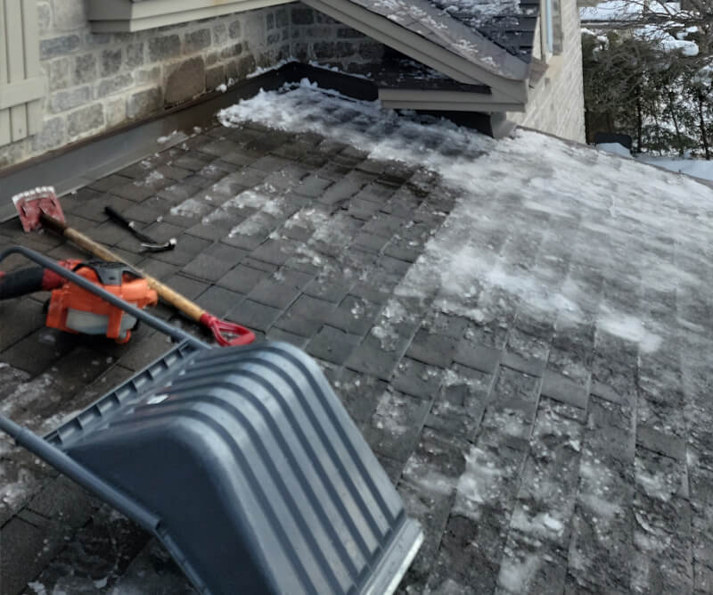 A snow-covered roof with patches of ice—ideal for ice dam removal in Manotick—a snow shovel and an orange ice chopper rest on the shingles near a brick house in winter.