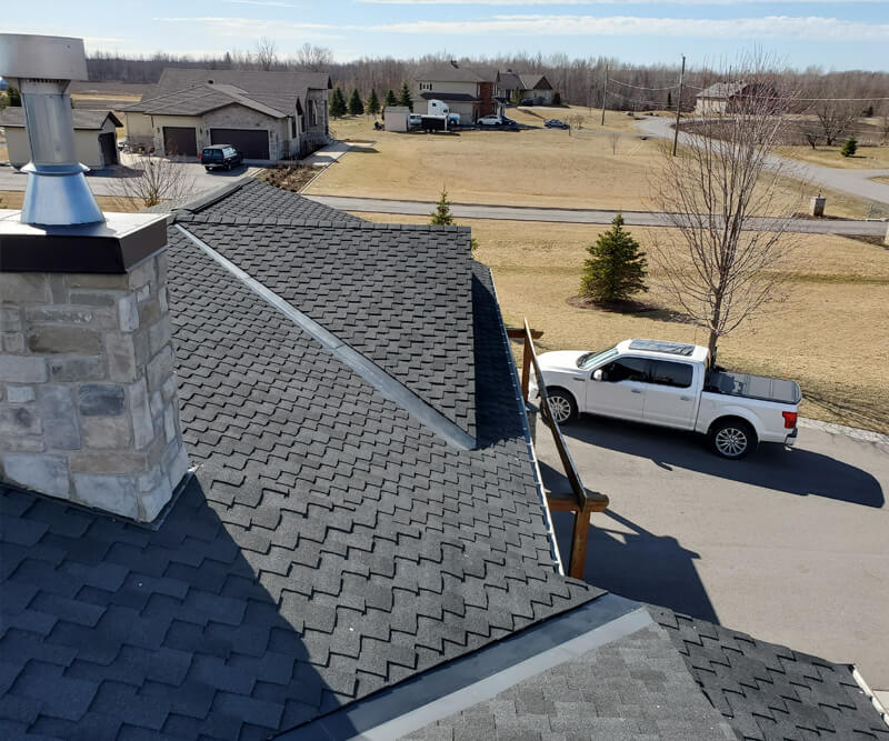 A white pickup truck is parked on a driveway beside a house with dark gray asphalt shingles, possibly after a roof replacement Ottawa homeowners might choose. The yard features dry grass, some small evergreens, and a leafless tree near the driveway.