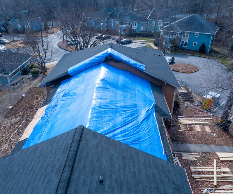 Aerial view of a house with a large blue tarp covering the roof, likely due to roof wind damage in Kanata, surrounded by trees and neighboring homes. Construction materials are visible on the ground near the building.