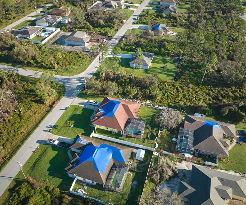 Aerial view of suburban houses in Kanata, Ontario, some with blue tarps covering storm-damaged roofs. Green lawns and trees surround the homes while roads divide the neighborhood.