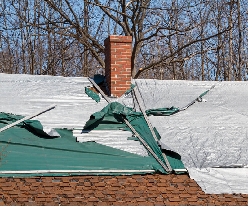 A Greely Ontario roof with damaged tarps flapping in the wind exposes shingles beneath. A brick chimney rises at the center, framed by leafless trees in the background.