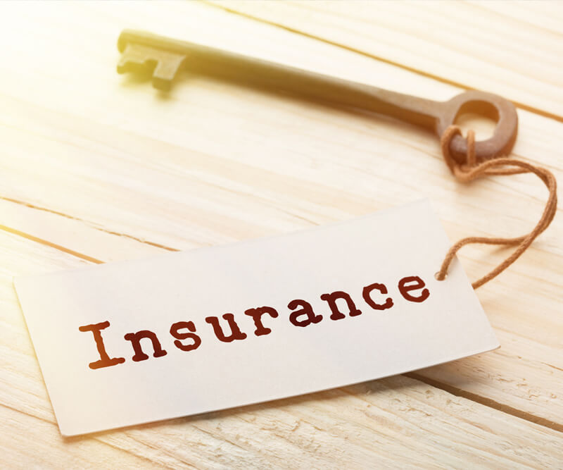 A vintage key rests on a wooden surface beside a white tag labeled Insurance in bold, brown letters. The tag, attached with string, hints at the importance of roof insurance Greely for safeguarding cherished possessions.