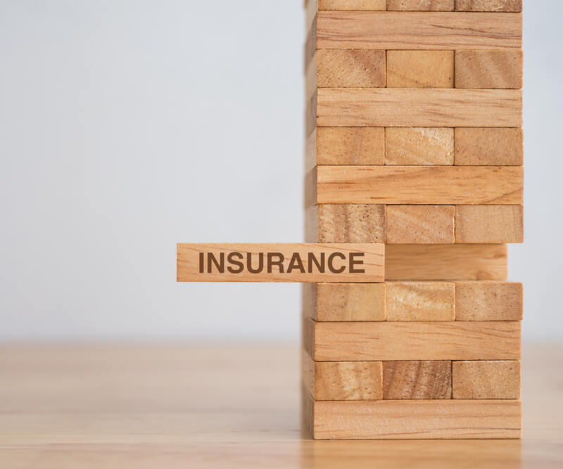 A Jenga tower made of wooden blocks, with one block labeled Greely insurance being pulled out, suggesting risk or instability without Greely insurance.
