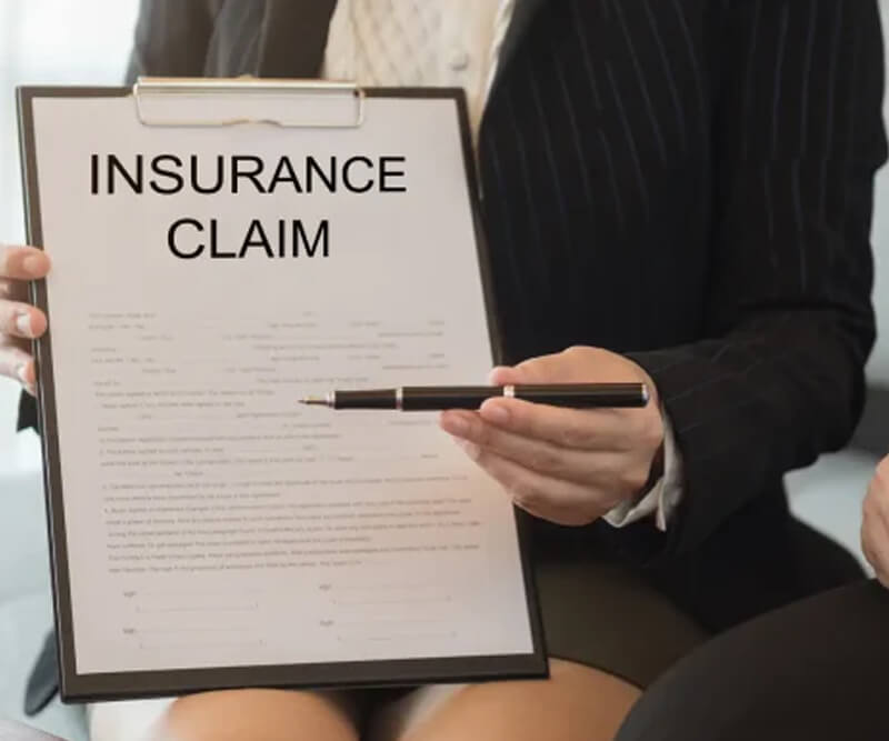 A person in business attire holds a clipboard with a document titled INSURANCE CLAIM, pointing to it with a pen, suggesting explanation or discussion about Greely insurance claims.