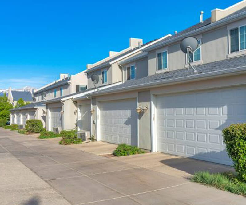 A row of modern, beige condos in The Glebe with double garages and satellite dishes on the roof, lined along a clean, paved driveway under a clear blue sky. Shrubs neatly border each unit.