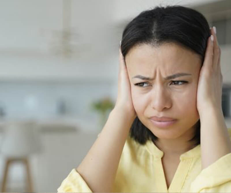 A woman in a yellow shirt covers her ears with her hands and looks upset, possibly reacting to loud roofing behaviours, sitting in a bright, blurry indoor setting.