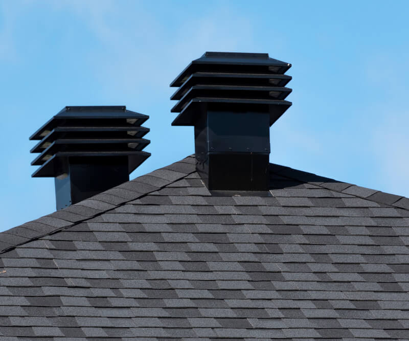 Two square black roof vents with horizontal slats on a gray shingled roof, set against a clear blue sky.