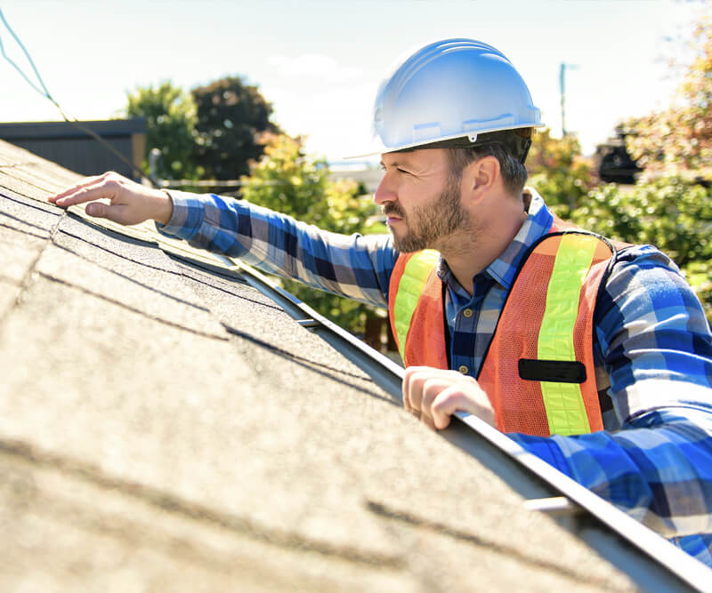 A construction worker from an Ottawa roofing company, wearing a hard hat, safety vest, and plaid shirt, inspects the shingles on a residential roof on a sunny day, surrounded by trees in the background.