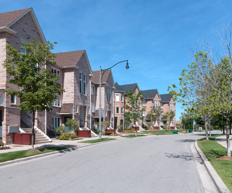 A quiet suburban street lined with modern, two-story brick houses featuring quality roofing in the Glebe, green lawns, and young trees under a clear blue sky.