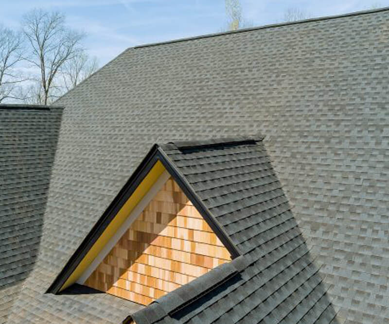 Close-up view of a gray shingled roofing Barrhaven home with a triangular dormer featuring tan wooden siding, set against a clear sky and bare tree branches in the background.