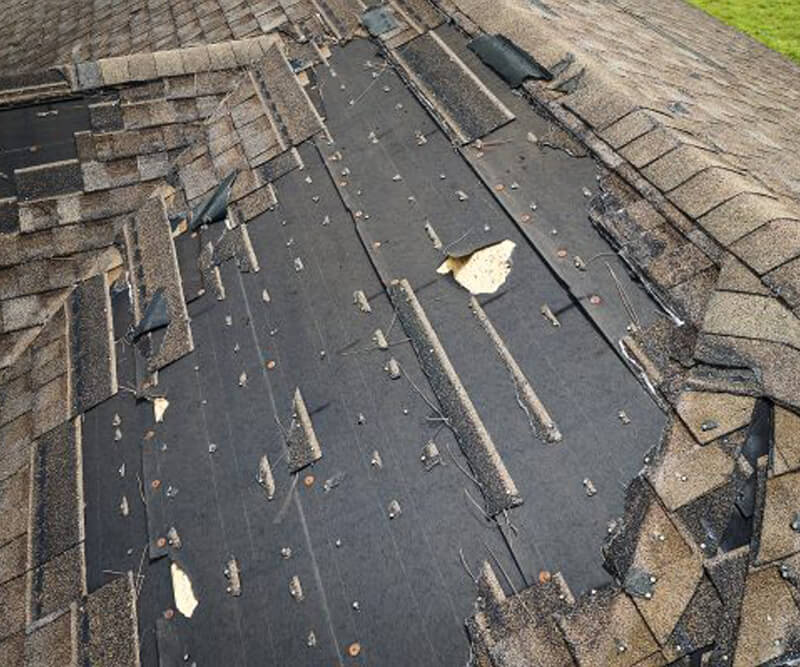 A roof with missing and damaged shingles exposes the black underlayment beneath. Some shingles are loose or curled up, and nails are visible, suggesting significant roof damage that may require a fiberglass roof replacement.