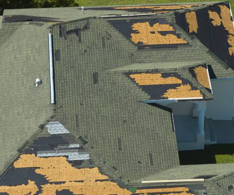 A house roof with missing and damaged shingles, exposing the underlying wooden sheathing in several large patches. The damage appears irregular and scattered across multiple sections—an issue best handled by Ottawa roofing experts.