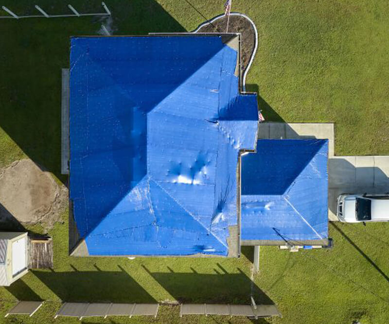 Aerial view of an Ottawa roof covered with a large blue tarp, surrounded by green grass, with a white car in the driveway. The tarp spans the entire roof, suggesting recent damage or repair work.