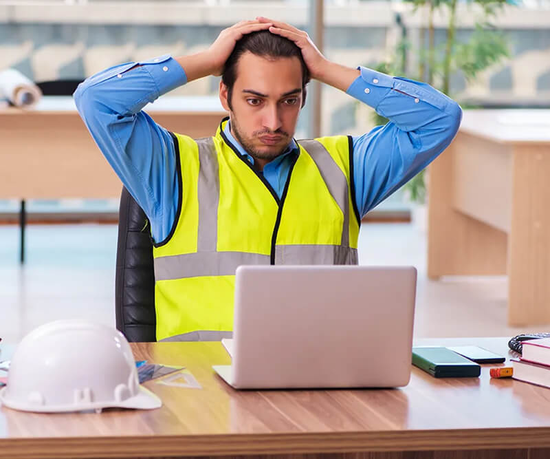 A man wearing a reflective safety vest sits at a desk, looking stressed with his hands on his head while staring at a laptop. A white hard hat and office supplies are on the desk in front of him, highlighting the challenges roofing experts face.