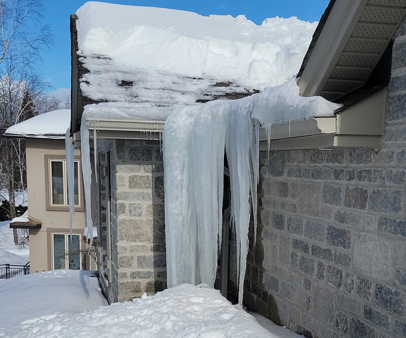 A large, thick sheet of ice hangs from the gutter of a house roof covered in snow, forming ice dams on the roof with icicles extending down to the ground. Snow piles up around the building on a bright winter day.