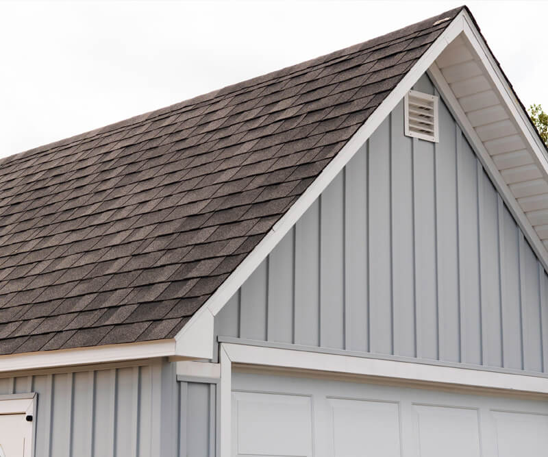 A close-up of a house with light blue vertical siding and a dark gray shingled roof, showcasing expert roof replacement Ottawa. There is a white vent near the roof peak and white trim along the edges.