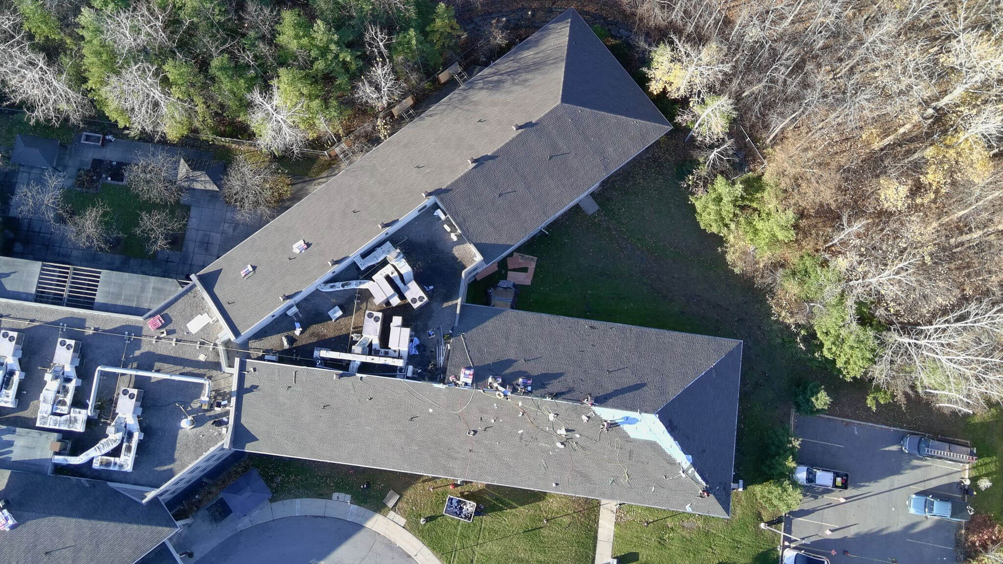 Aerial view of a large building with workers and materials on the roof, showcasing active commercial roofing in Ottawa. The site is surrounded by trees, grass, and parking areas, with construction activity visible on the rooftop.