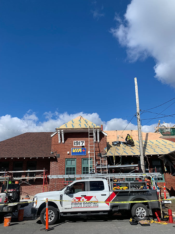 A brick train station with a 1917 VIA sign is being re-roofed. Workers and ladders are on the roof, and a Firon Roofing truck with equipment is parked in front under a blue sky with scattered clouds.