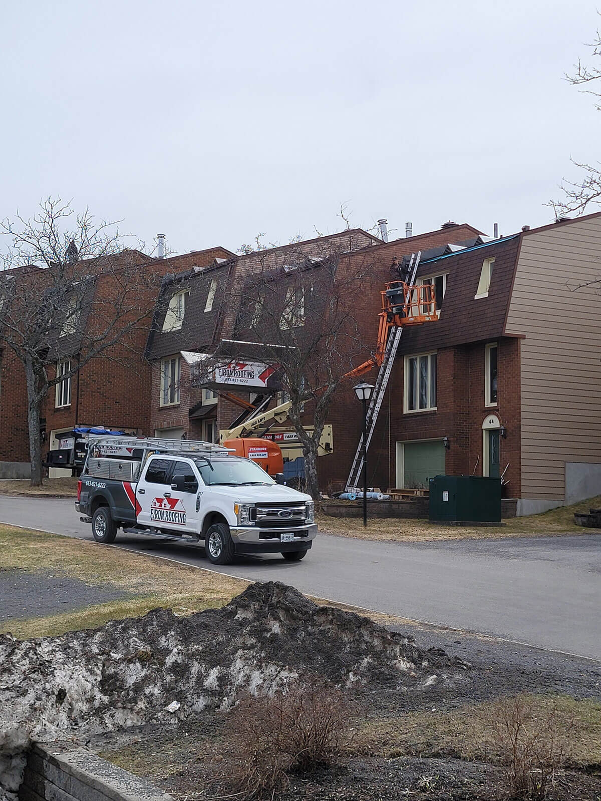 Workers on an elevated lift repair the roof of a multi-story brick building, exemplifying professional roofing for business. Nearby, a truck and equipment with Les Couvreurs Duro-Toit branding are parked amid snow and bare trees in early spring or late winter.