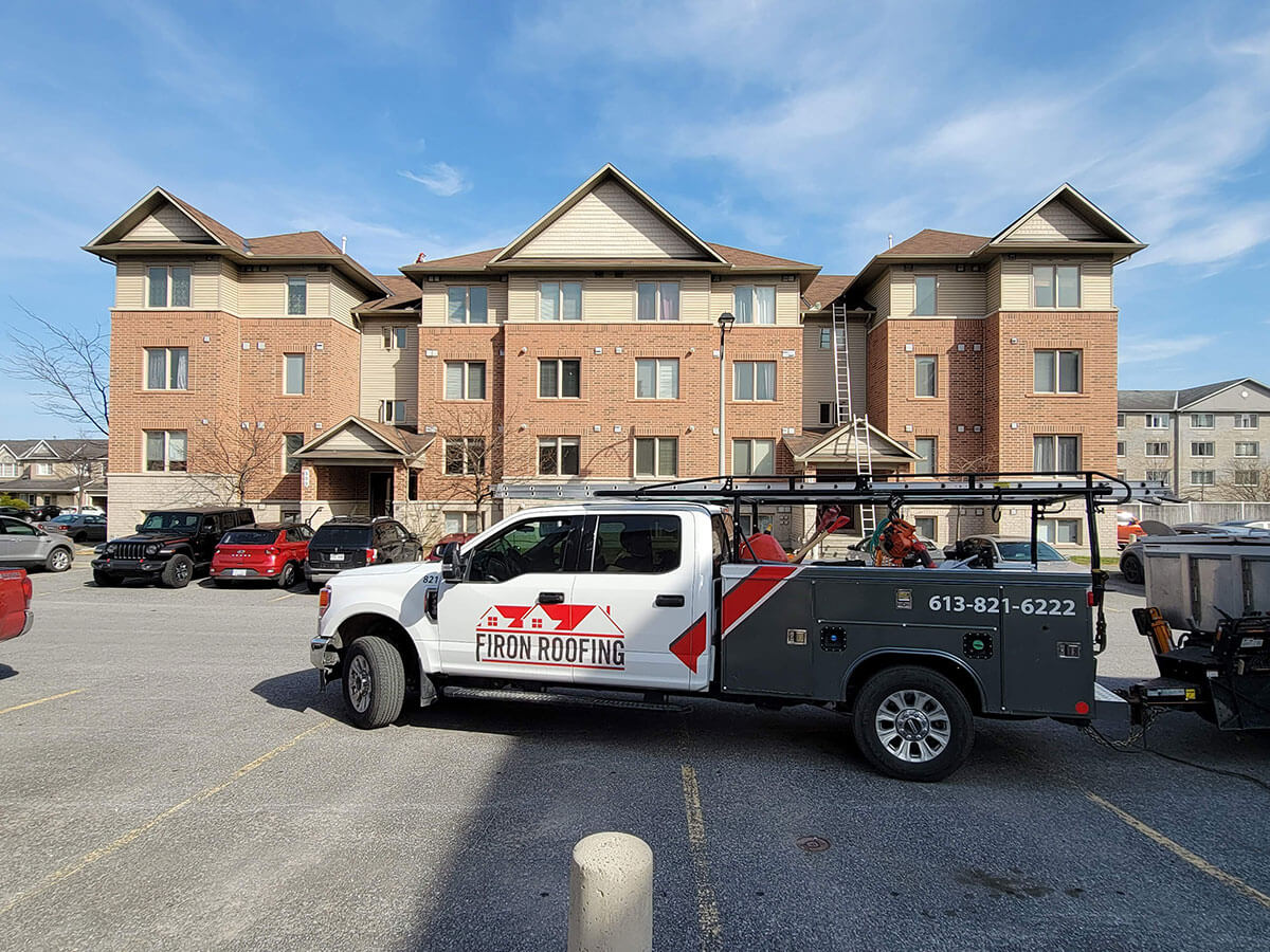 A pickup truck with Firon Roofing branding, highlighting their B2B solutions, is parked in front of a three-story brick apartment building. A ladder leans against the building, with several other cars nearby beneath a clear blue sky.