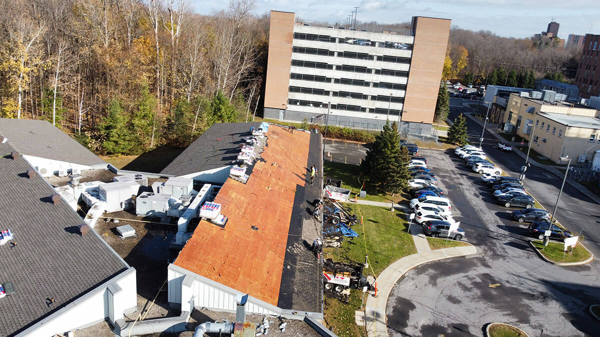 Aerial view of workers replacing the roof of a building, with commercial roofing Ottawa materials laid out. Nearby are parked cars, trees with autumn leaves, and a multi-story parking garage in the background.