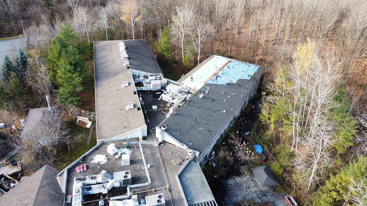 Aerial view of a large building with roof damage and debris scattered around. Part of the roof appears to be missing or under repair. The building is surrounded by trees, some with autumn leaves, and a cleared area is visible nearby.
