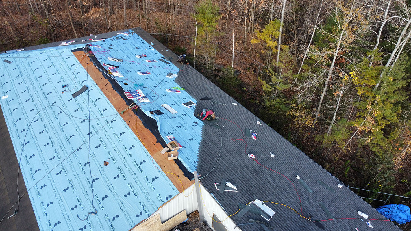 Aerial view of a house roof under repair by Firon Roofing Gatineau, with blue underlayment partially installed and roofing materials scattered. Surrounding area has trees with autumn foliage.